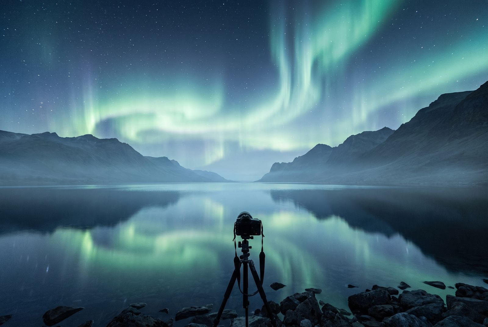 Northern lights over a still mountain lake, a camera on a tripod at the shore